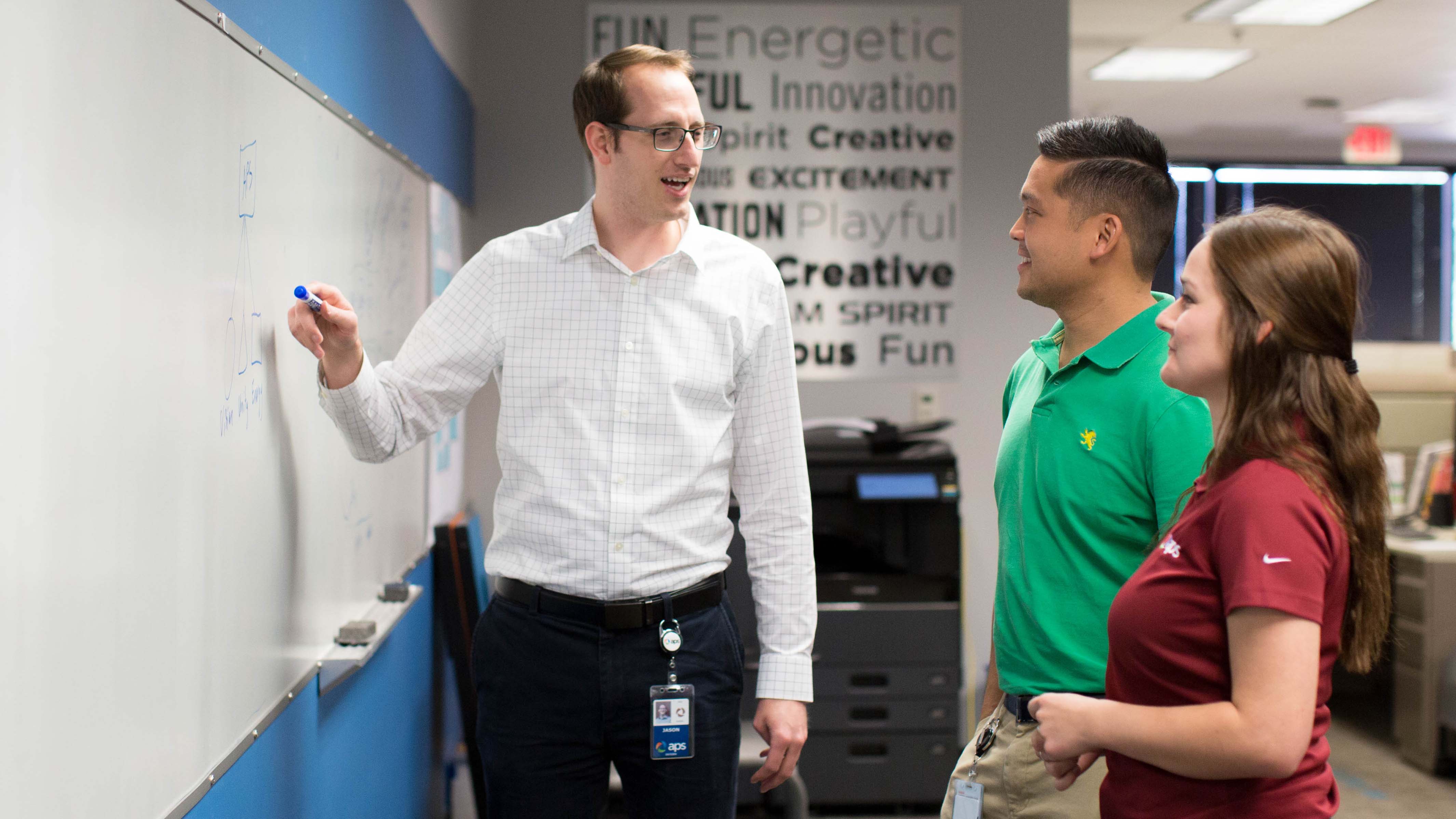 Three APS interns standing at a whiteboard discussing what is displayed on the whiteboard