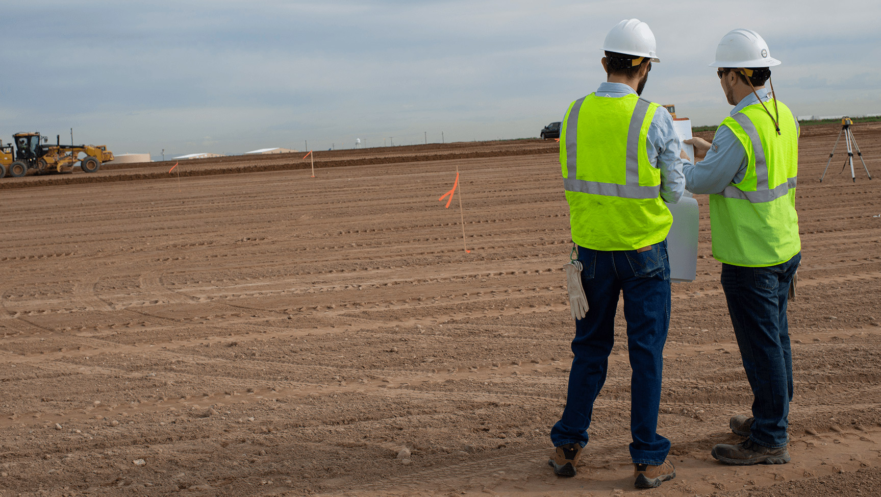 Two construction workers looking at a document on a dirt lot.