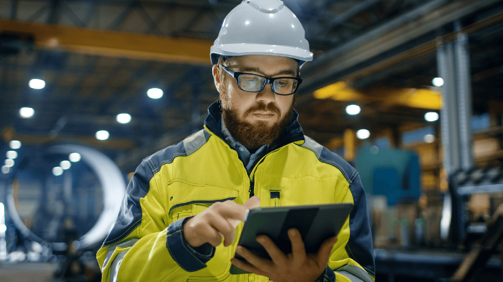 Man in a warehouse facility wearing a hard hat looking at a tablet.