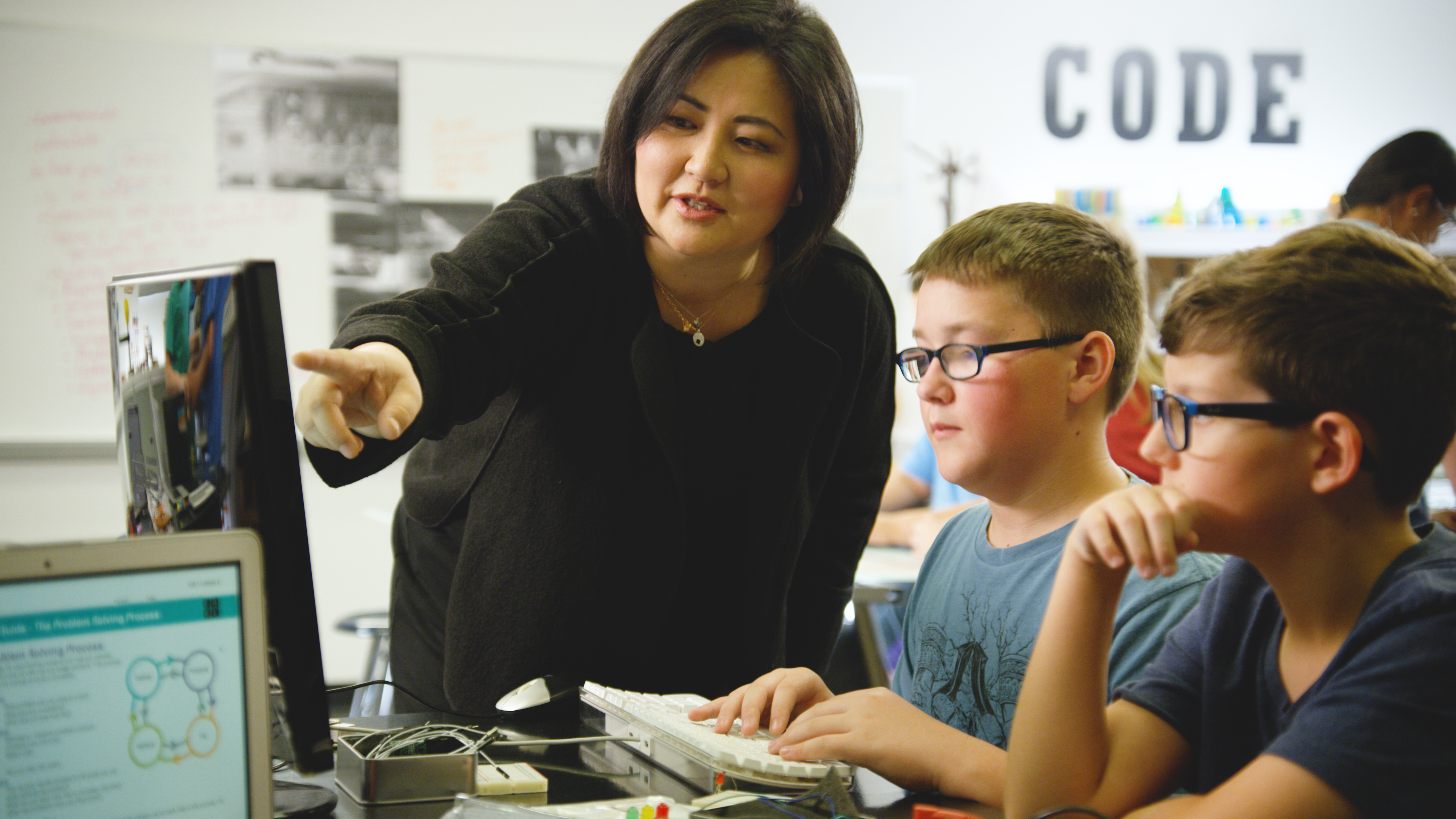 A teacher assisting students in a classroom pointing to the computer monitor at a desk