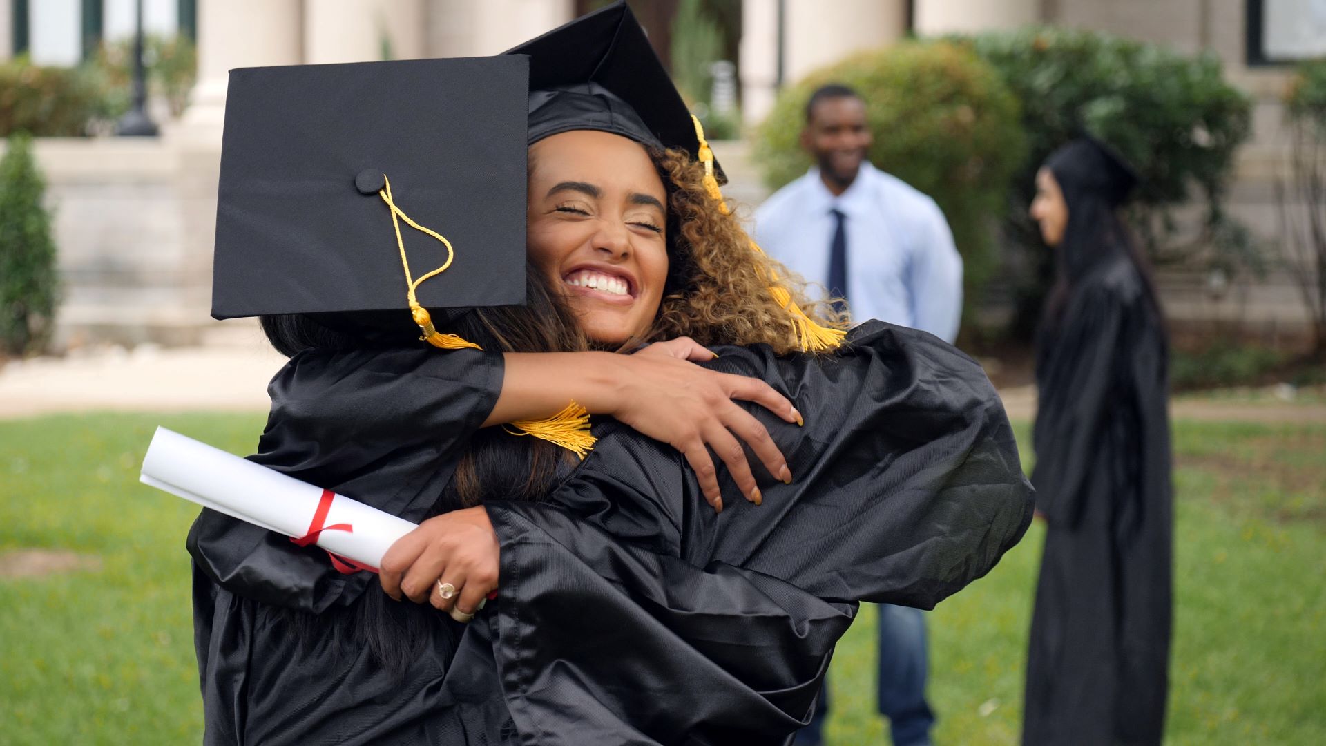 Woman with graduation gown and cap embracing another graduate