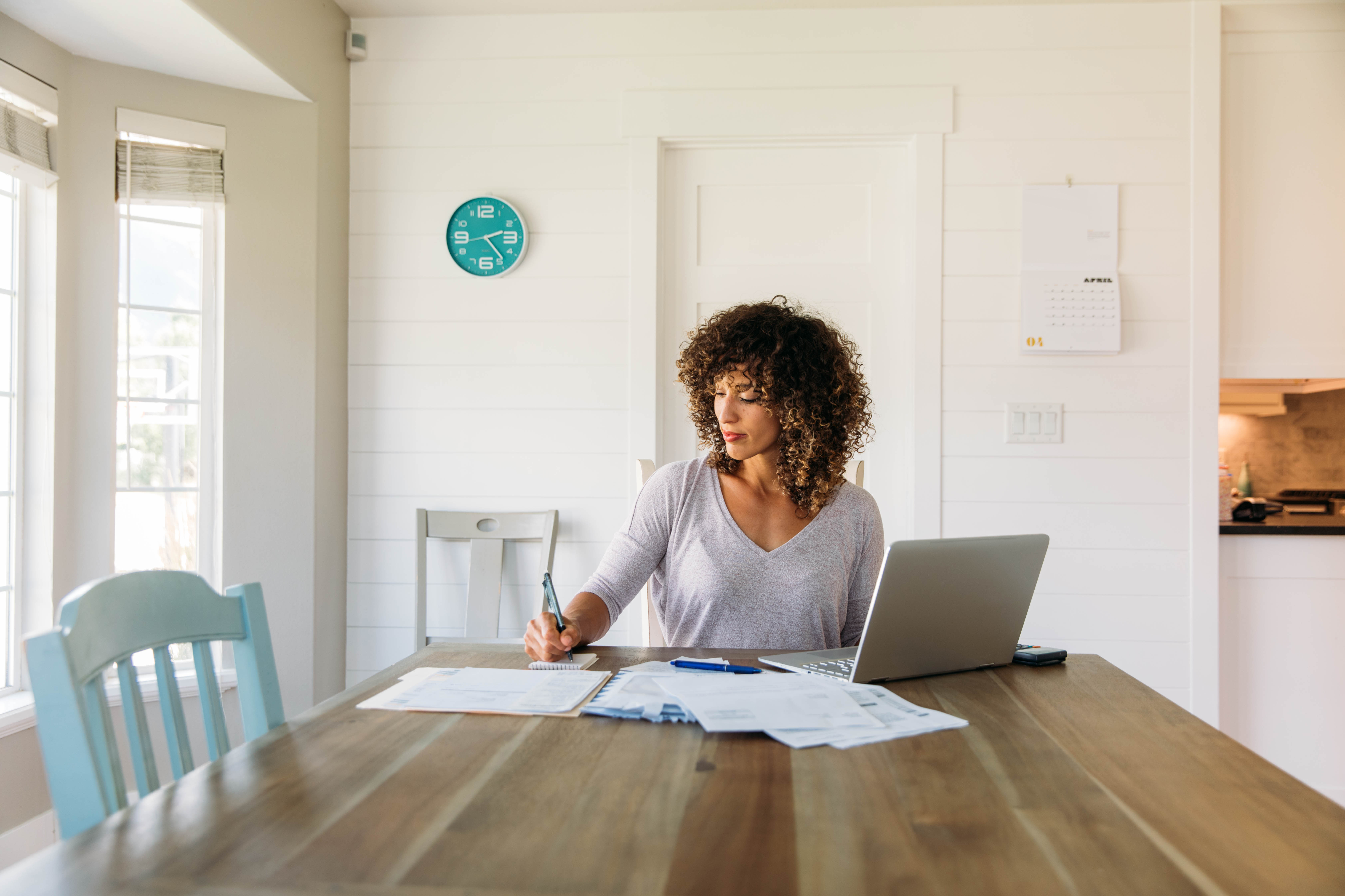 A woman sitting at the end of a table working on some paperwork with an open laptop nearby.