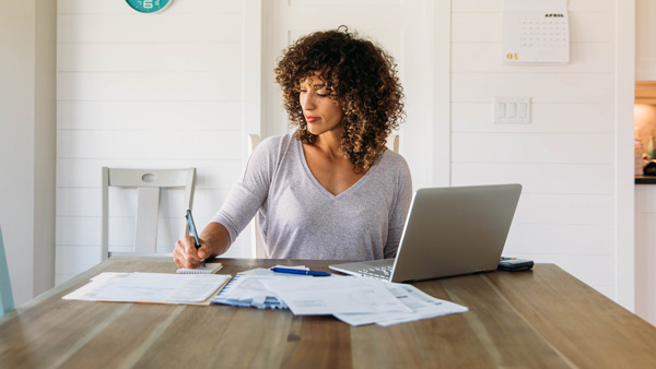 A woman sitting at the end of a table working on some paperwork with an open laptop nearby.