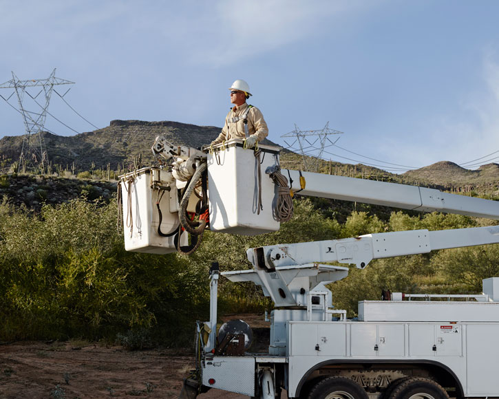A color photograph showing APS Employee Jason Adams wearing a white hard hat, safety glasses, and a protective harness standing inside the white bucket of an extended utility boom truck. Behind him is a desert landscape featuring green brush, saguaro cacti, rolling mountains, and tall steel electrical transmission towers under a clear sky.