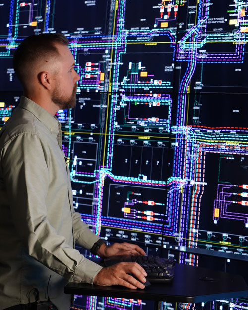 A color photograph of a man with a beard standing in a darkened control room. He is operating a computer mouse and keyboard at a standing desk, looking straight ahead at a massive wall of glowing monitors. The screens display a highly detailed, complex schematic of an electrical grid mapped out in bright neon lines and nodes.