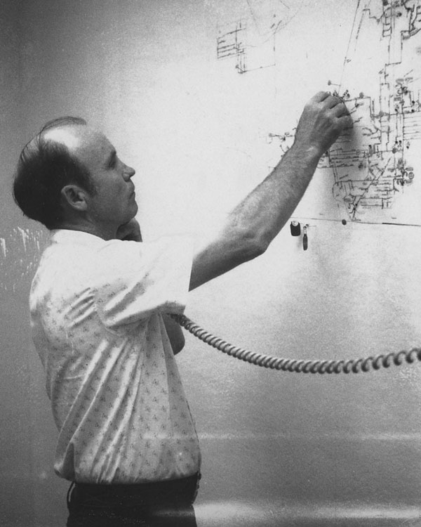 A black and white photograph of a man working in an office. He is looking at a large, intricate paper map taped to the wall. He is holding a coiled-cord telephone receiver to his ear with his shoulder while using his right hand to point at or adjust a marker on the map.