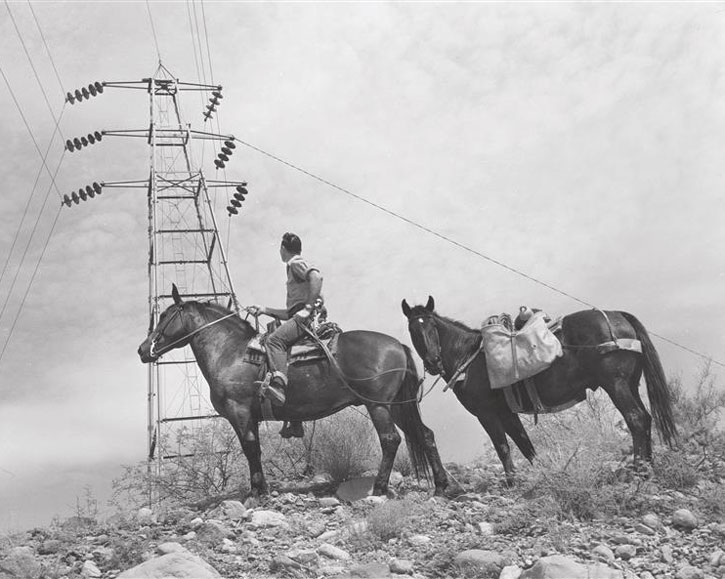 A black and white photograph showing a man riding a horse across rocky, scrub-filled terrain. He is looking over his shoulder toward a second horse trailing behind him, which is loaded with canvas pack bags. To the left, dominating the background, is a tall metal lattice electrical transmission tower with heavy power lines stretching across a partly cloudy sky.