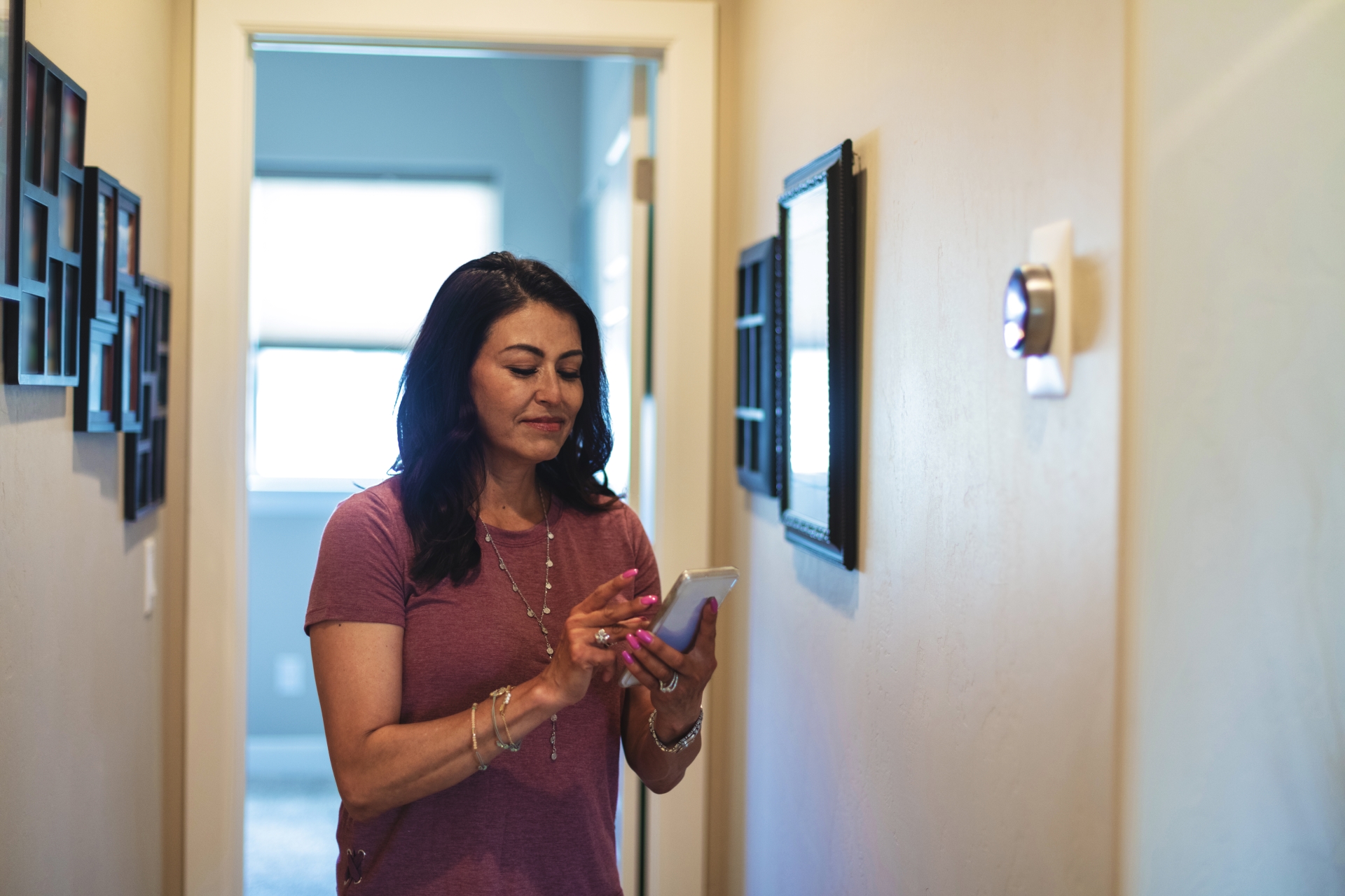 Woman standing in the hallway looking at her phone of her home next to a smart thermostat