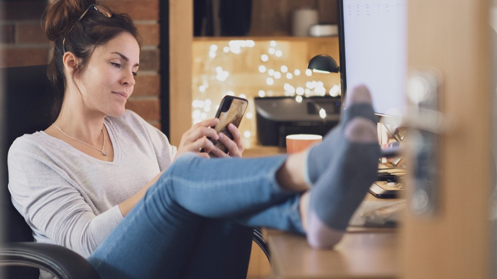 Woman looking at her phone in home office with her feet up on the desk.
