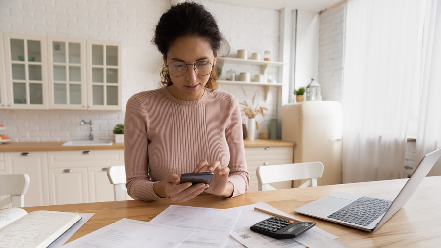 Woman sitting at a kitchen table with a laptop, calculator and papers typing on the calculator 