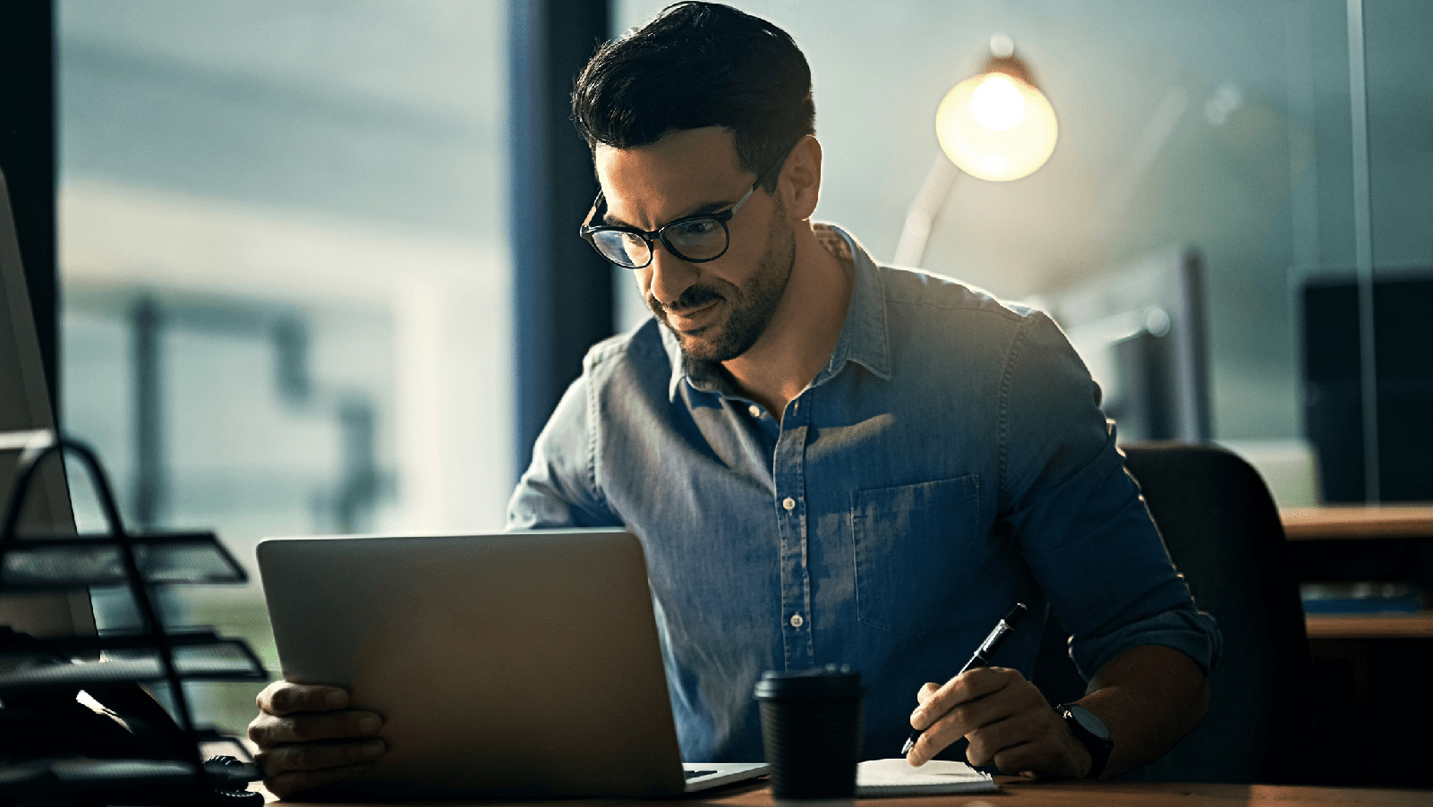 Man wearing glasses and working on a laptop at his desk.
