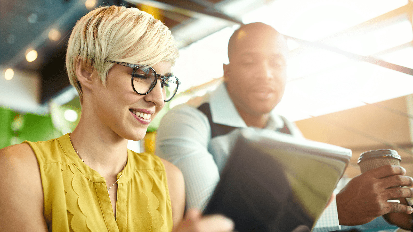 Woman in glasses and man holding coffee looking at a tablet.