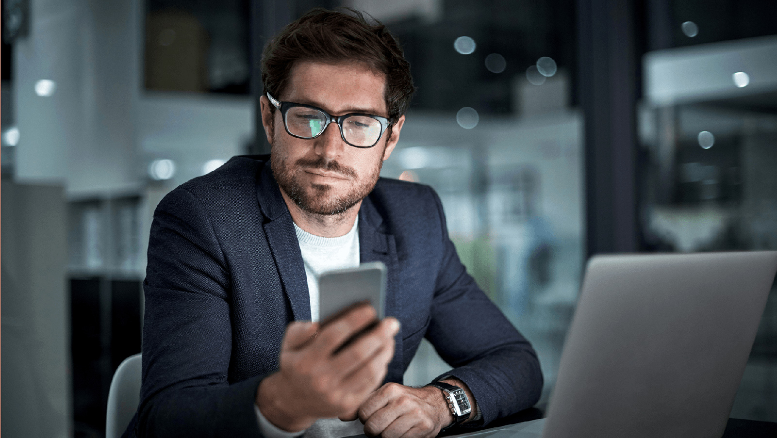 Man in glasses looking at his phone at a desk.