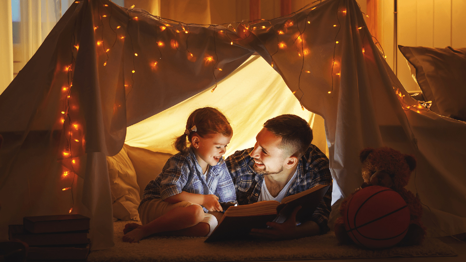 Father reading to his daughter in a blanket fort.