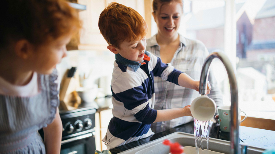 young boy pouring water into the kitchen sink with his mother watching