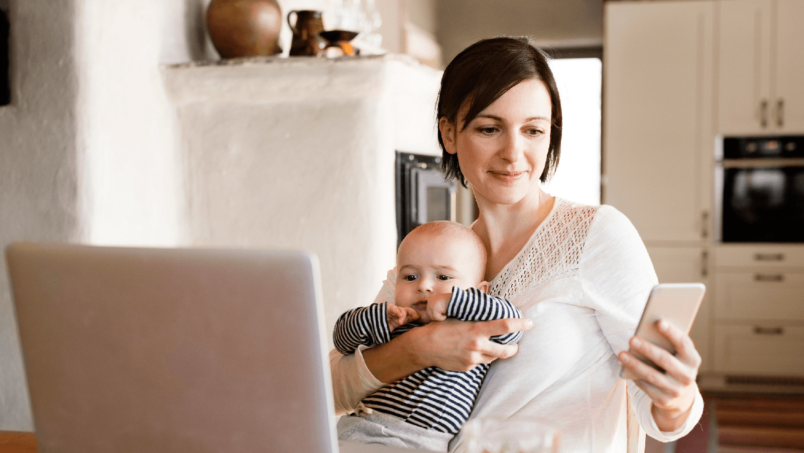 Woman smiling and holding a baby while looking at her phone in the kitchen.